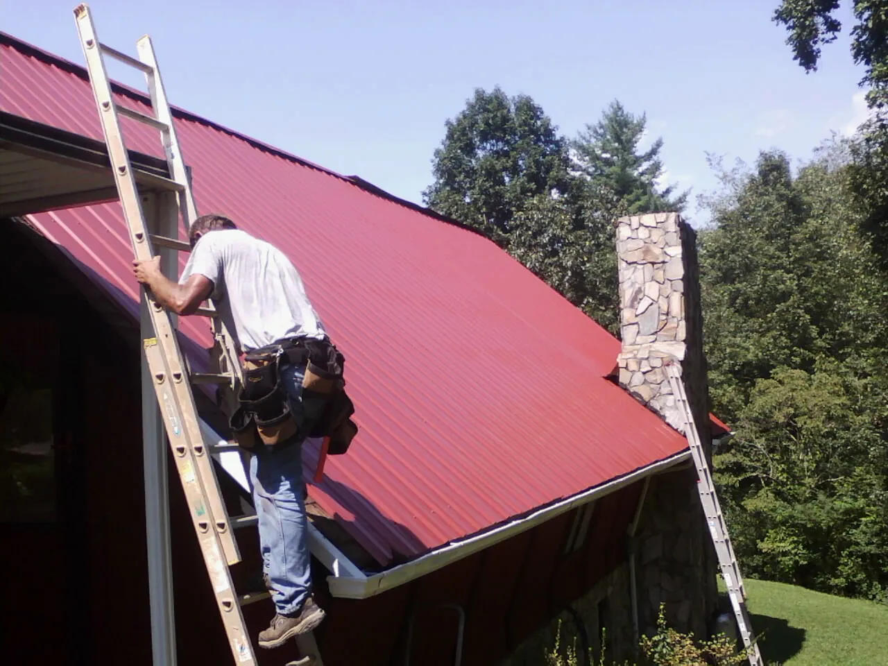 Roofer on a ladder beside a red metal roof and stone chimney — Soffit & Fascia Repair by Roofing Brothers in Coraopolis